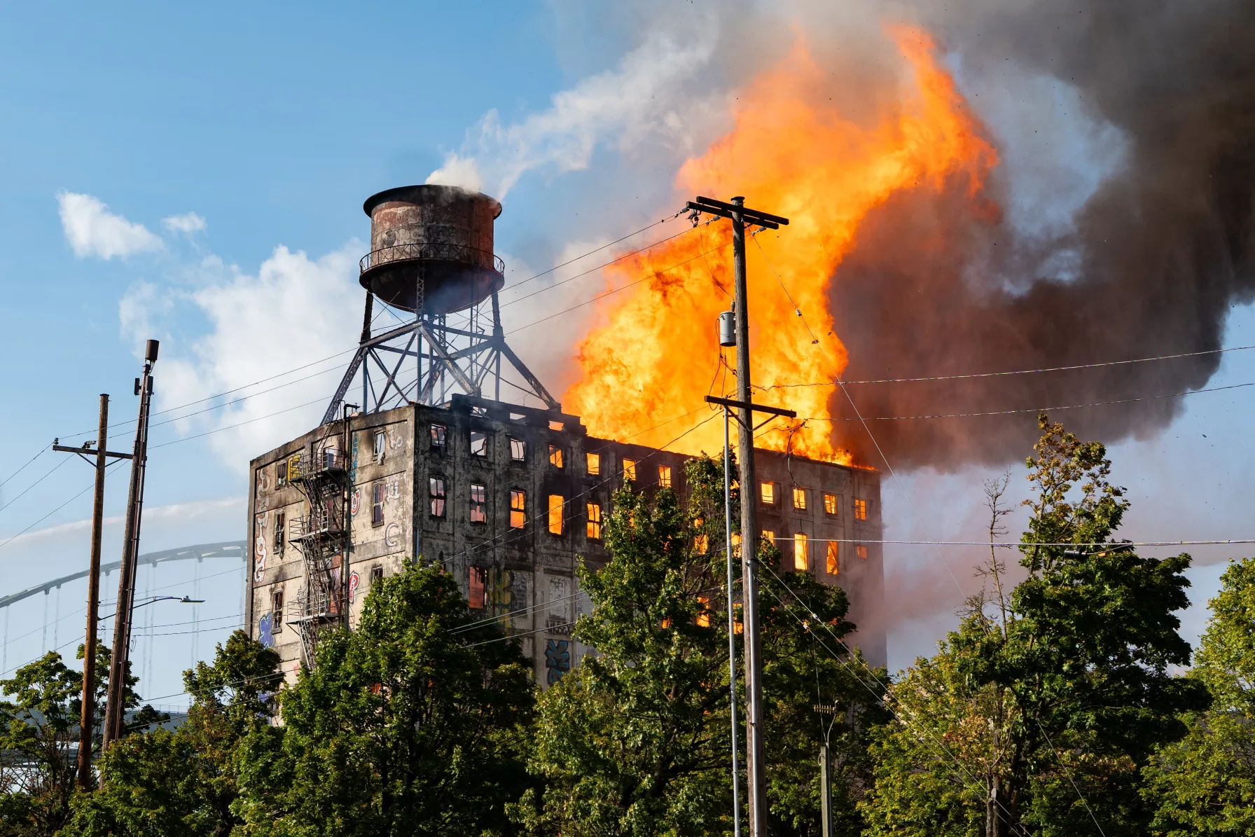 Flames and thick smoke pour from the multi-story Centennial Mills warehouse in Northwest Portland as its rooftop water tower stands over the blaze, with the St. Johns Bridge visible in the background.