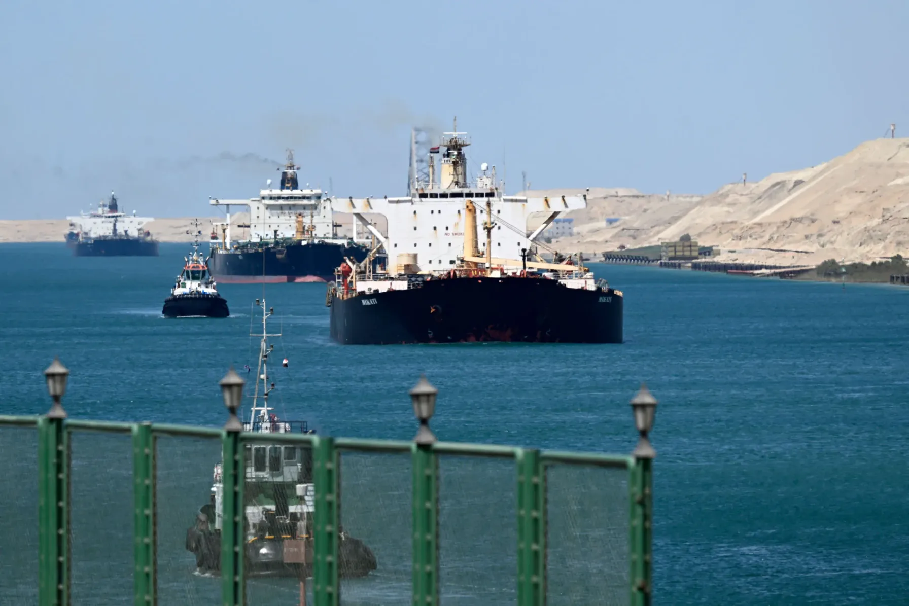 Oil tankers and tugboats transiting a desert waterway chokepoint with sand dunes on either bank