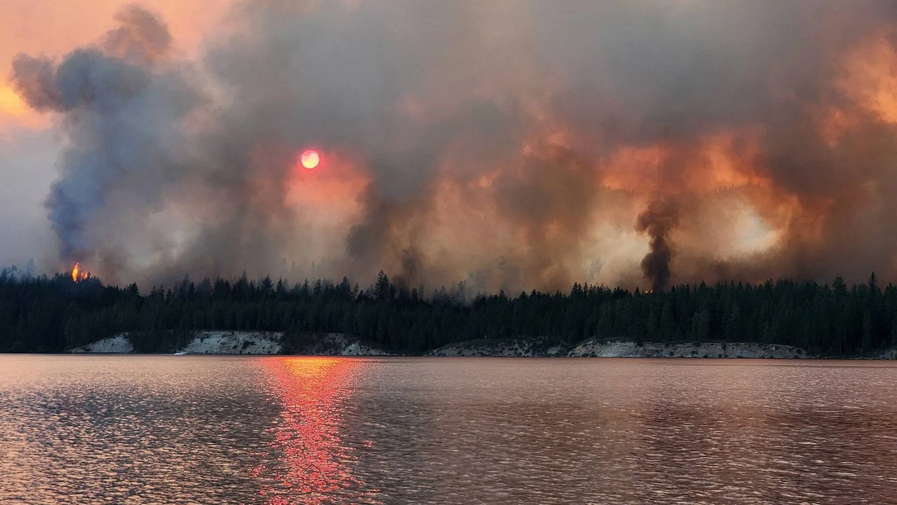 Heavy smoke and flames from the Kaiser Canyon Fire rising over trees near a lake in Eastern Washington