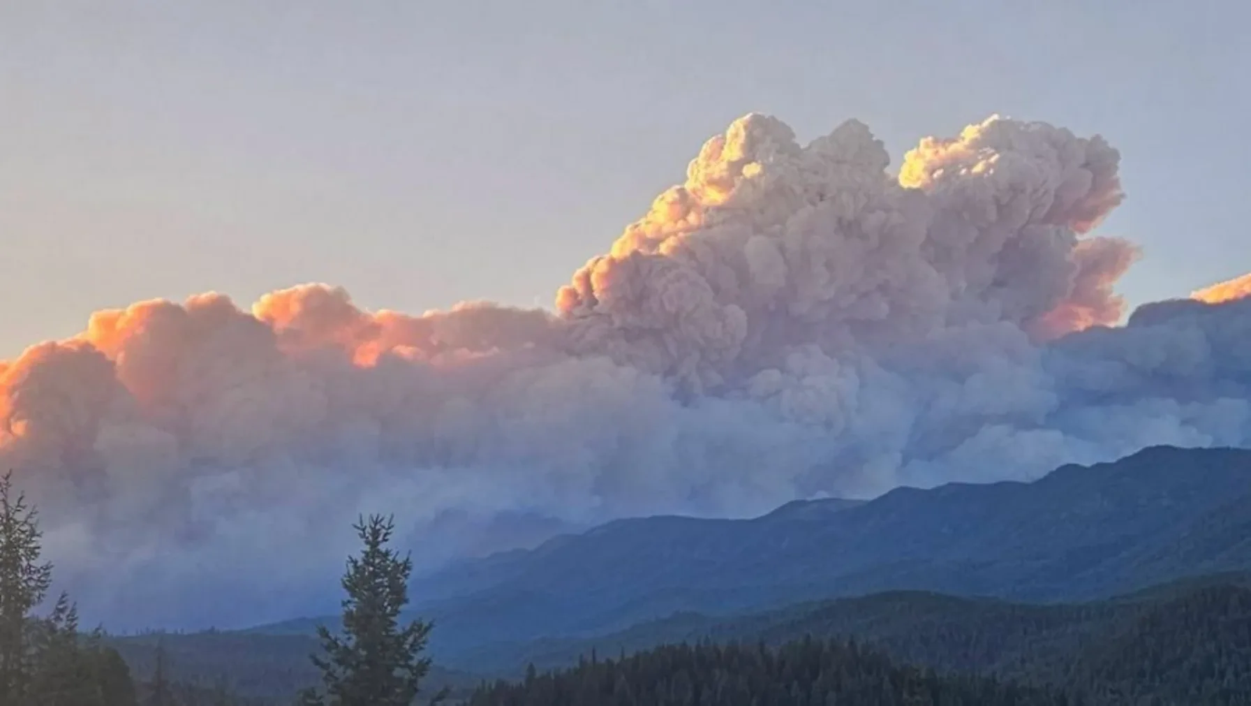 A massive orange-lit pyrocumulus smoke plume rises over forested mountains at sunset from the Little Giant Fire in Chelan County, Washington.