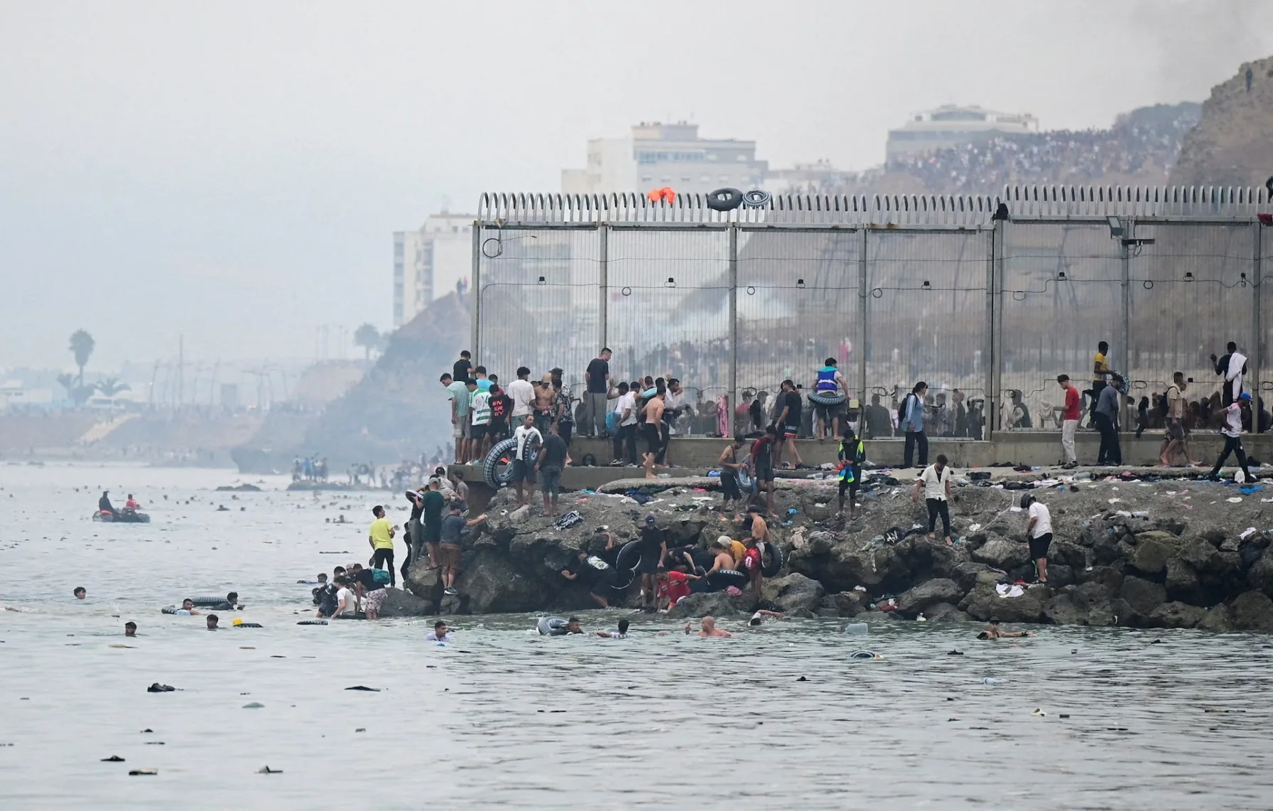 Migrants swimming and climbing along a breakwater and border fence at Ceuta, with people gathered on the rocks and in the water near the fence