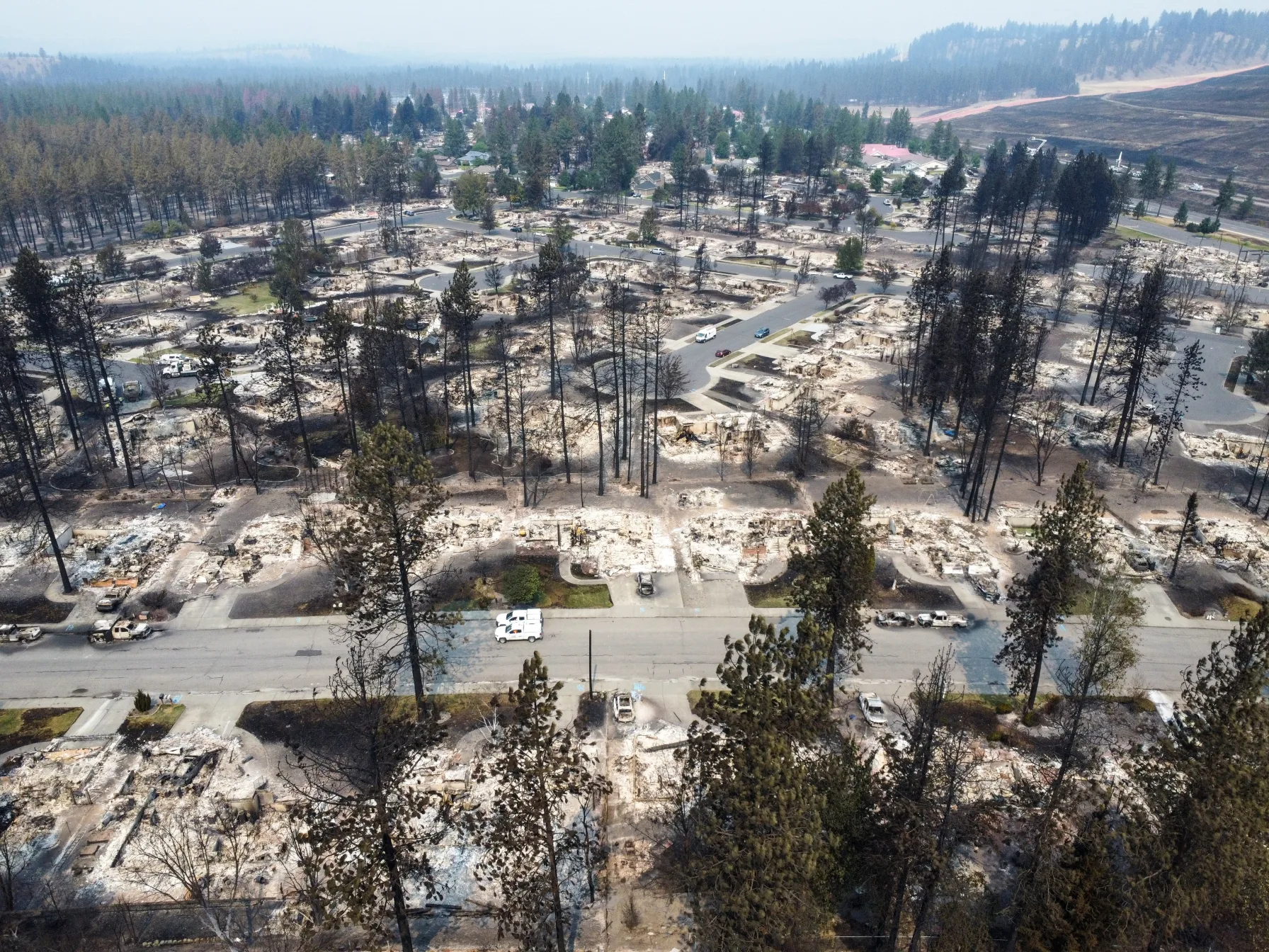 Aerial view of a Spokane neighborhood reduced to ash and building foundations after the Old Trails Fire, with vehicles working the streets amid smoke haze