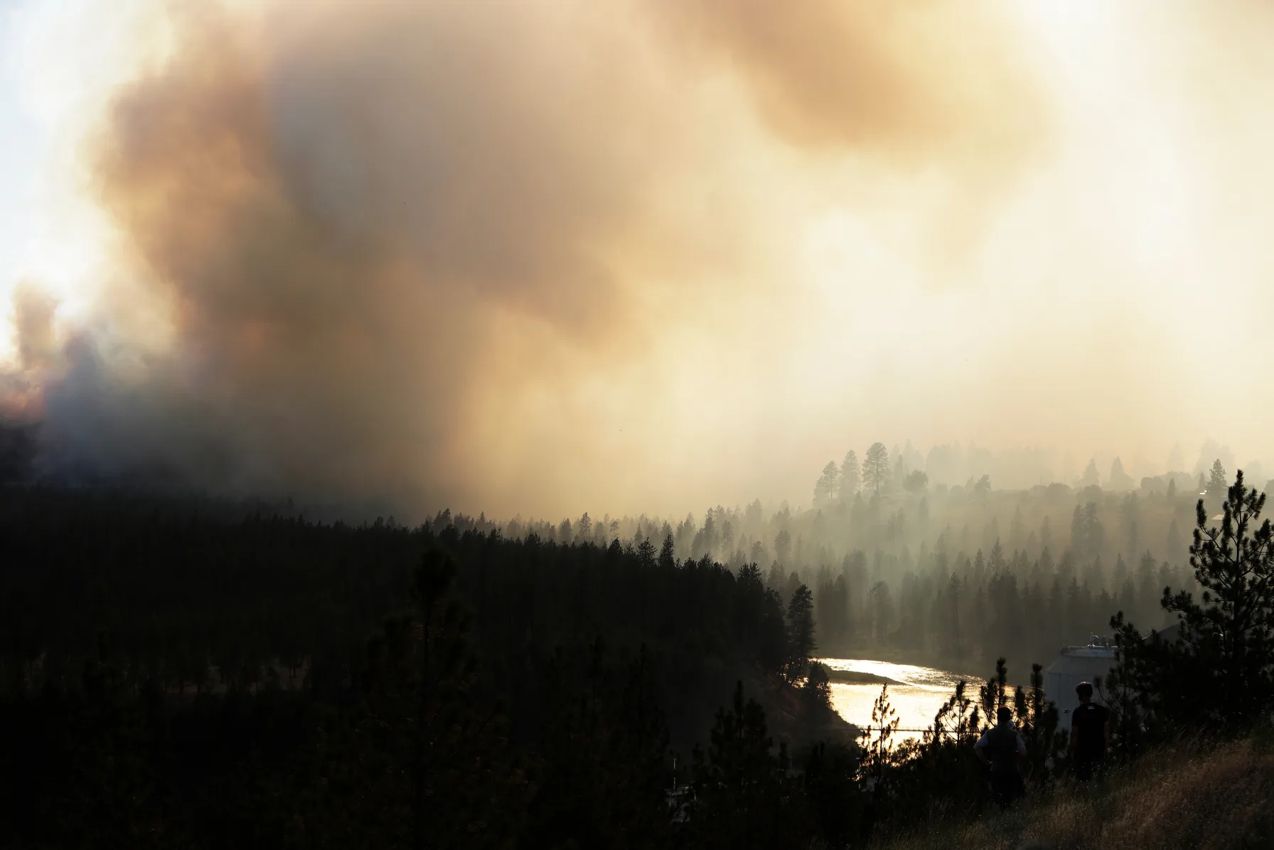 Thick wildfire smoke rises over the Spokane River near the Old Trails Fire as two onlookers watch from a ridge, August 1, 2026