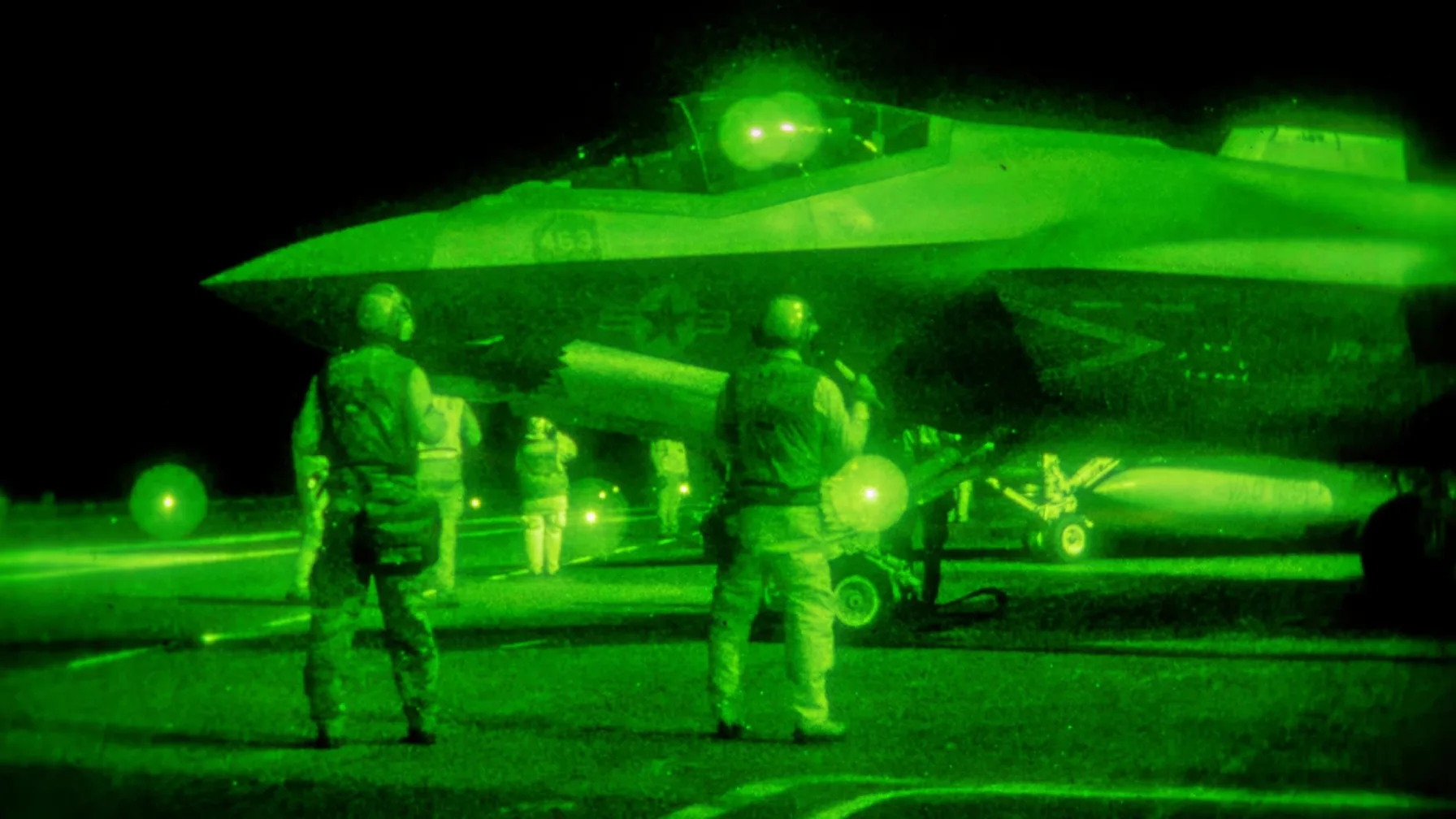 Night-vision view of U.S. Navy personnel on a flight deck with a fighter jet during carrier operations