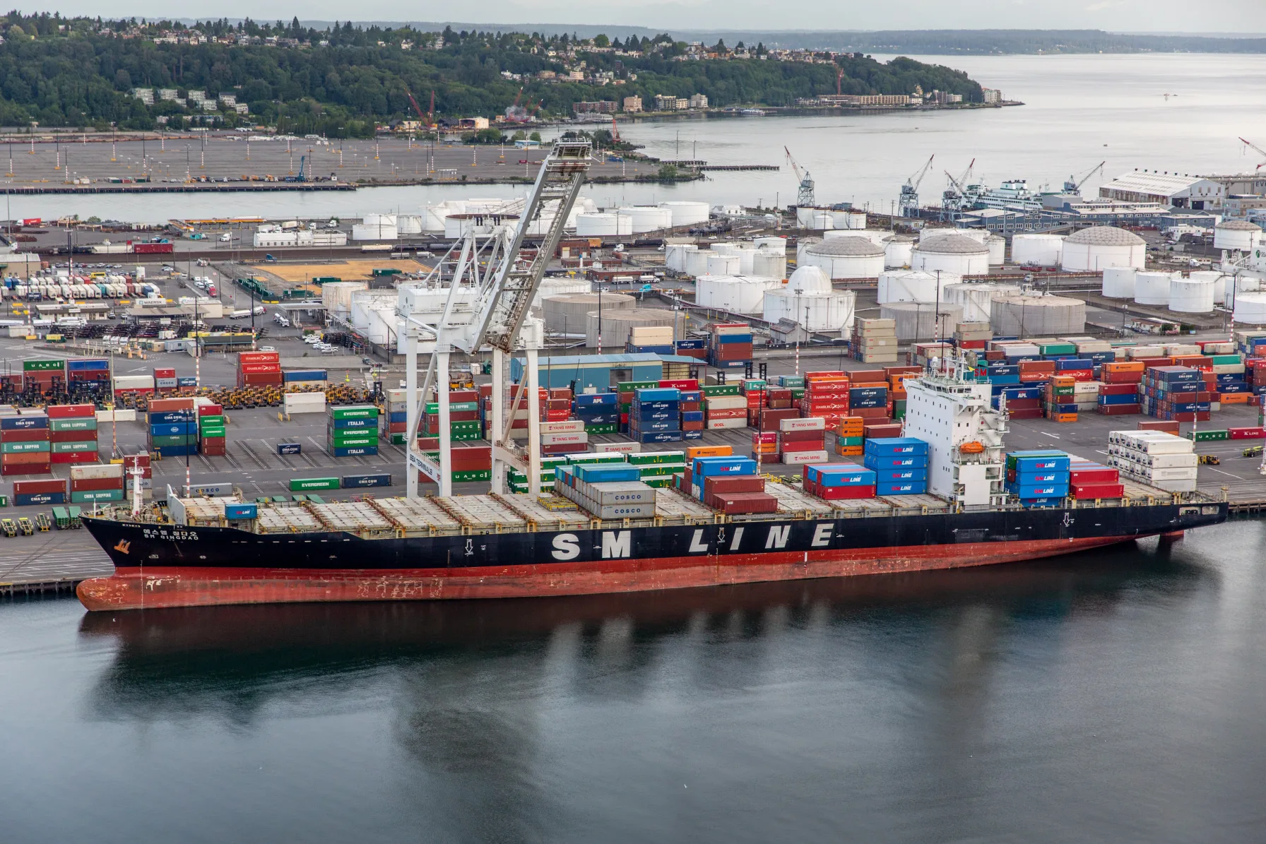 A container ship docked at the Port of Seattle with shipping containers stacked on the dock and the Seattle skyline in the background